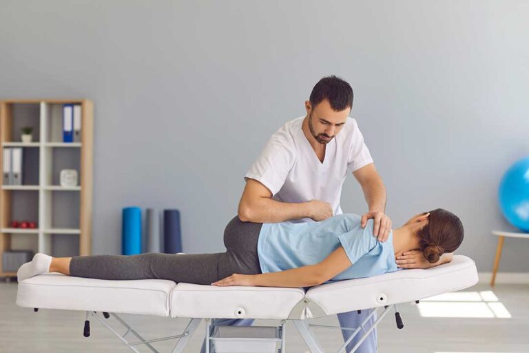 Person gently stretching a tight shoulder beside a window, with a massage ball and anatomy chart nearby.